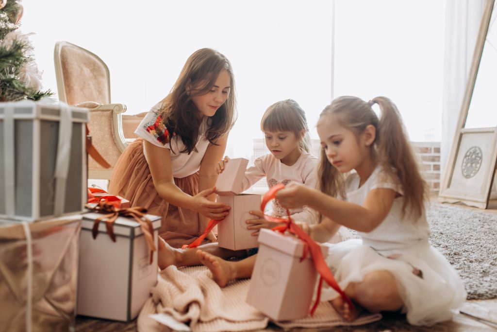 Happy young mother and her two charming daughters in nice dresses sit on the carpet and open New Year's gifts in the light cozy room